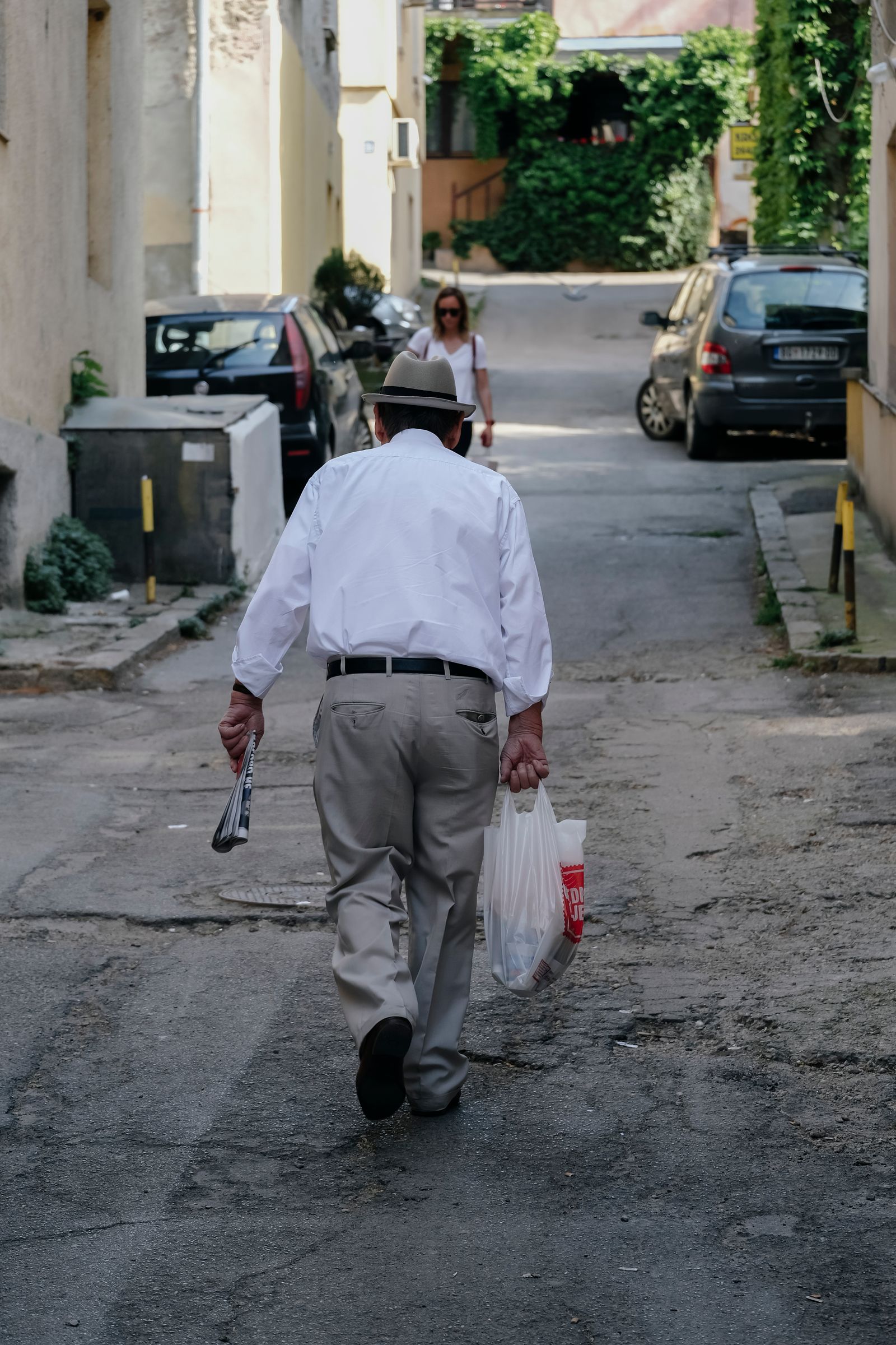 An older man in a white shirt and a pale fedora walks away from the camera up a sunlit Mediterranean side street, a folded newspaper in one hand and a shopping bag in the other