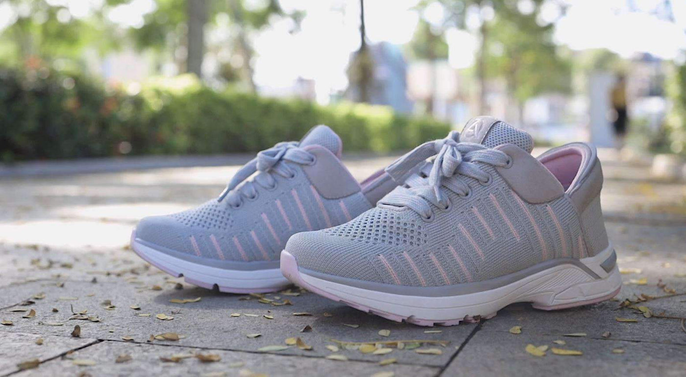 A pair of Zeba Rose Gray hands-free sneakers in grey knit with pink stitching, photographed on a paved path